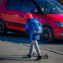 Gutt med sekk og sparkesykkel foran rød bil